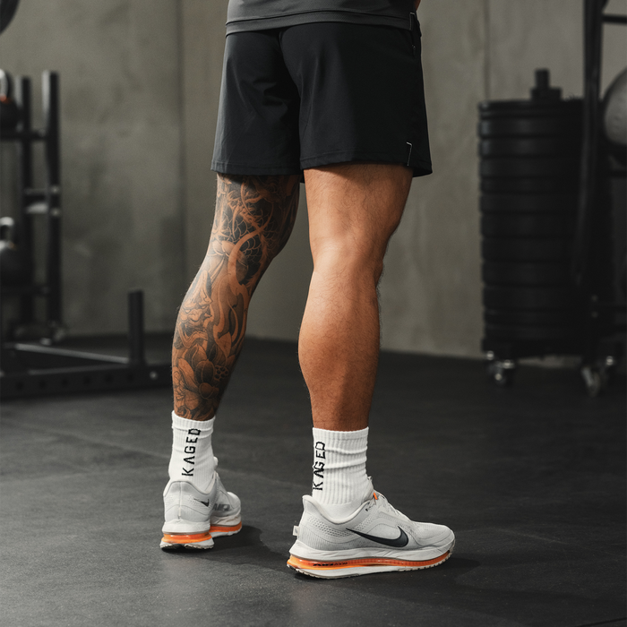 Rear view of Ascent Performance Socks in white on a model, showing ribbed crew height and KAGED branding on the cuff in a gym