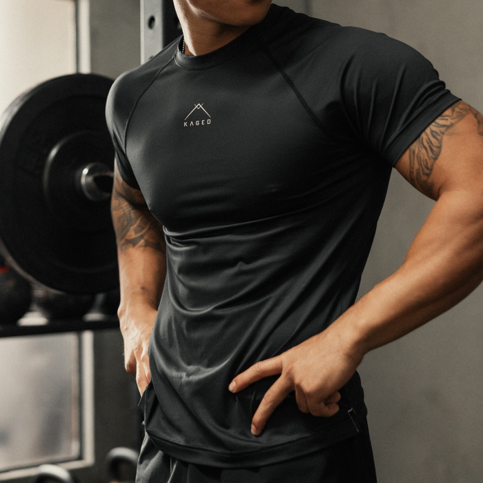 Front view of black Ascent Performance Tee showcasing athletic fit and moisture-wicking 4-way stretch fabric with gym equipment background