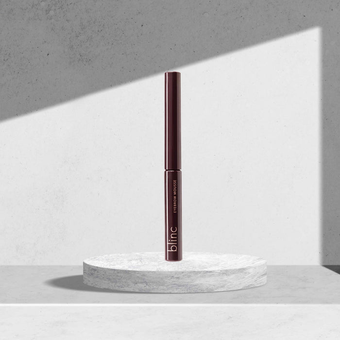 Eyebrow Mousse | Blinc in a sleek dark brown tube standing upright on a round marble platform with soft natural light and a minimalist gray background.