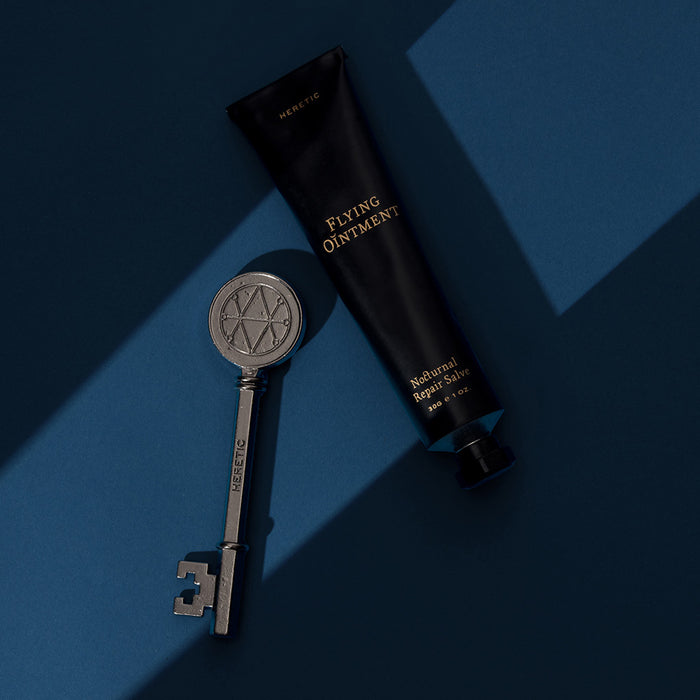 A black squeeze tube labeled 'FLYING OINTMENT' and a matching silver decorative key on a dark blue background with geometric shadows.