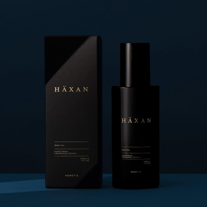 A matte black bottle of HÄXAN body oil stands next to its matching box packaging on a dark navy blue background.