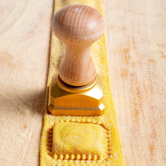 A rectangular brass pasta stamp with a wooden handle, featuring the text 'LA GONDELA' on the stamp surface stamping out pasta on a wooden table.
