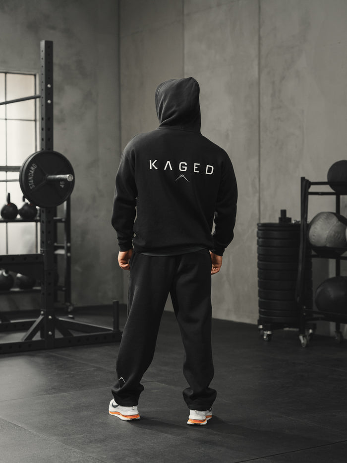 Rear view of Ascent Hoodie with hood up, showing large KAGED logo on back and relaxed athletic fit on model in gym setting