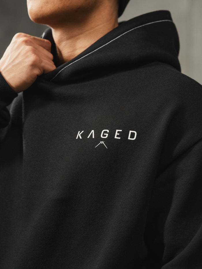 Close-up of Ascent Hoodie showing white KAGED chest logo and contrast hood stitching on model with neutral gray background
