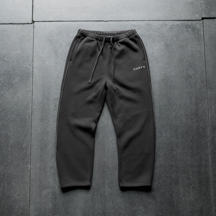 Flat lay of Ascent straight-leg sweatpants showcasing black midweight fleece, drawstring waistband, white side-seam stitching, and Kaged logo on gray tiled floor