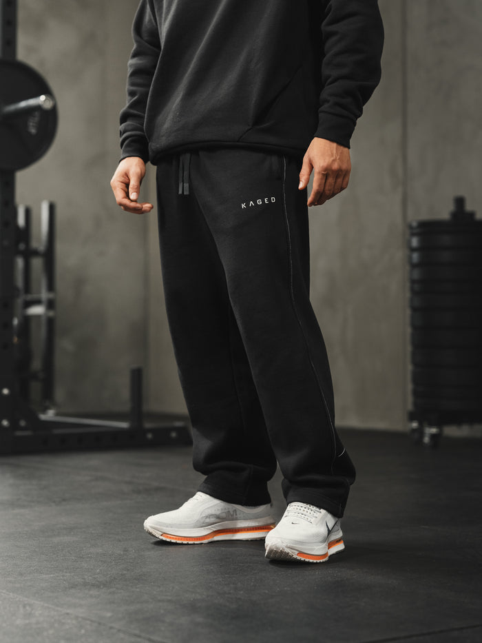 Front view of black Ascent straight-leg sweatpants showing white side-seam stitching, Kaged logo on left thigh, drawstring waistband, gym setting