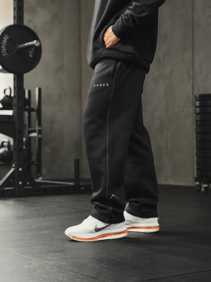 Side view of Ascent straight-leg sweats in black showing white side-seam stitching, KAGED logo and Ascent ankle symbol in a gym setting