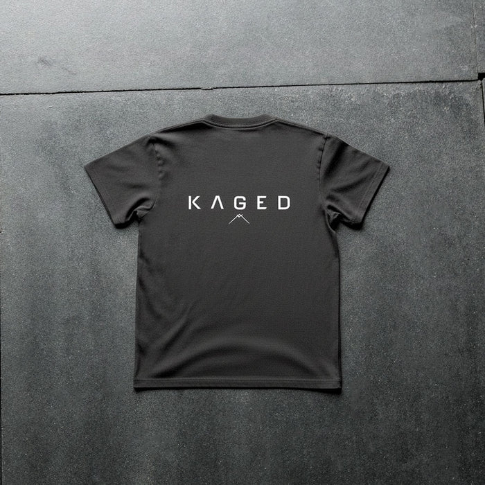 Back view of Ascent Tee showing large white KAGED wordmark and small mountain logo on a black tee against a dark textured floor