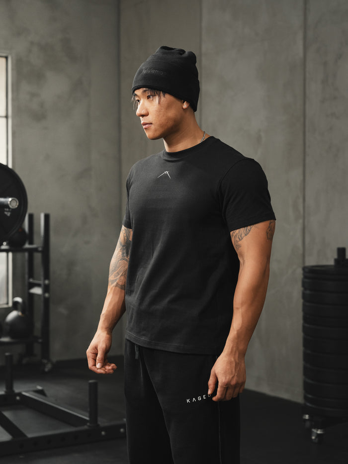 Model wearing fitted black Ascent tee standing in a gym with weight rack and plates in the background