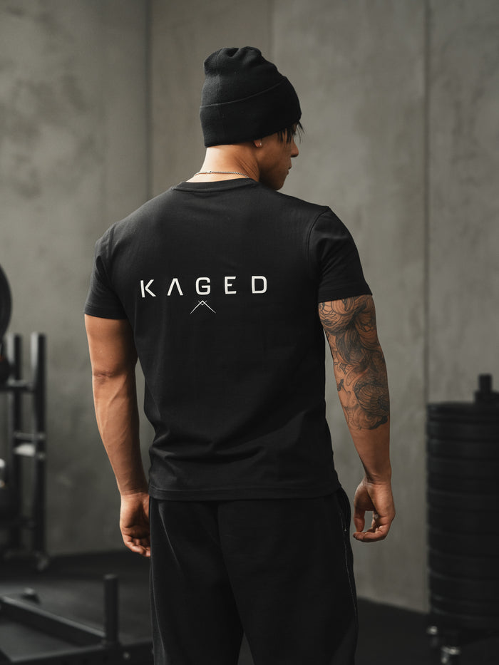Rear view of black Ascent Tee on model showcasing large KAGED wordmark on back and fitted silhouette in a gym setting