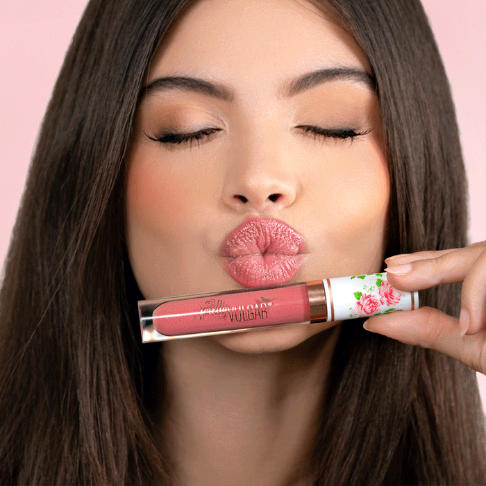 A woman with long brown hair puckers her lips in a kiss gesture while holding a tube of CEO of Lip Gloss | Pretty Vulgar in a soft pink shade with floral packaging against a light pink background.