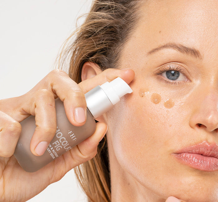 Close-up of a woman applying Chocolate Fig Vitamin Recovery Serum | Farmhouse Fresh to her cheek, demonstrating skincare routine with serum drops on her skin.