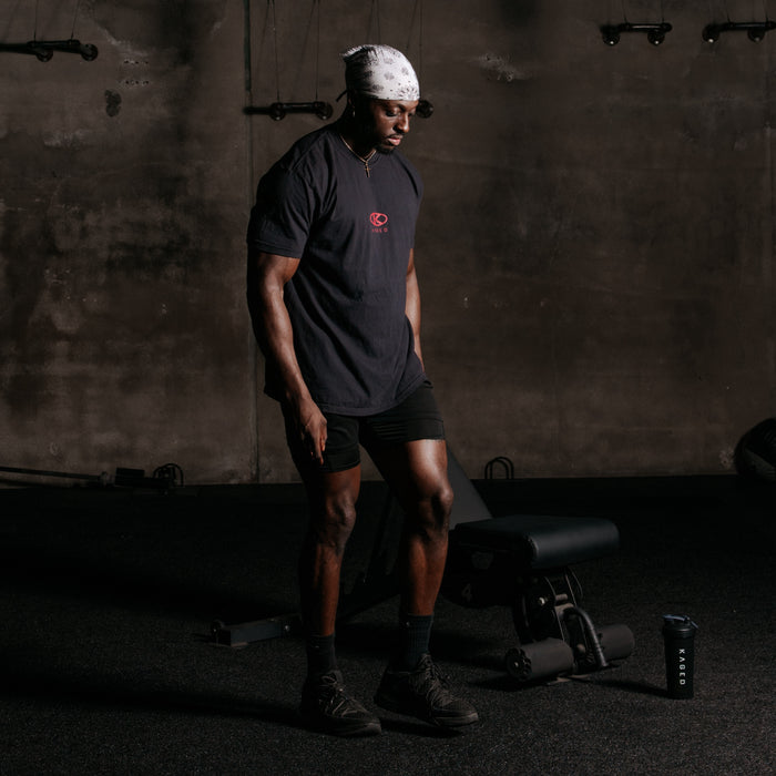Three-quarter view of Circle Tee showcasing red KAGED circle logo on a black short-sleeve tee in a dim gym
