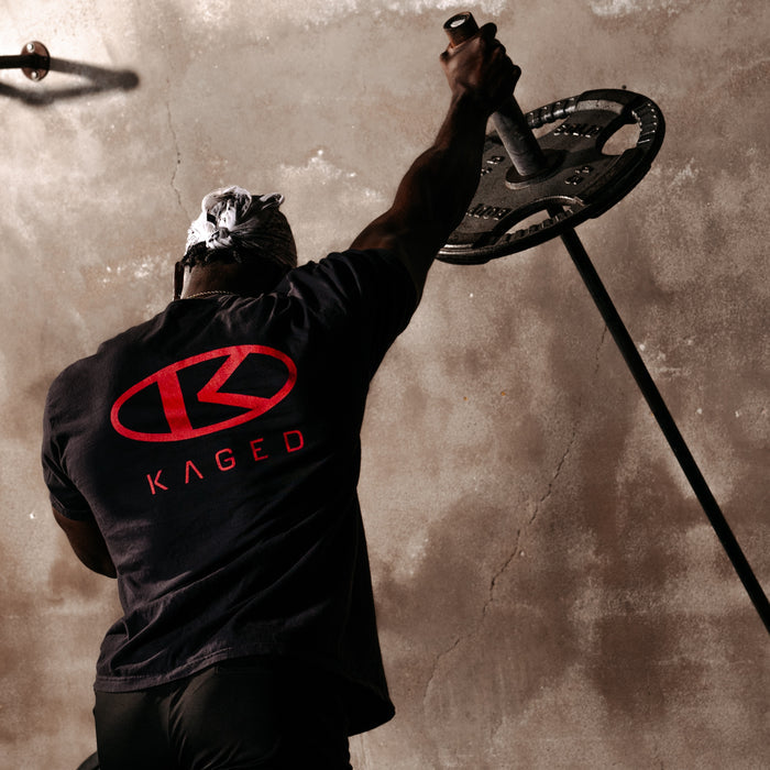 Rear view of Circle Tee showing red KAGED circle logo on the back as a model lifts a barbell against a concrete wall