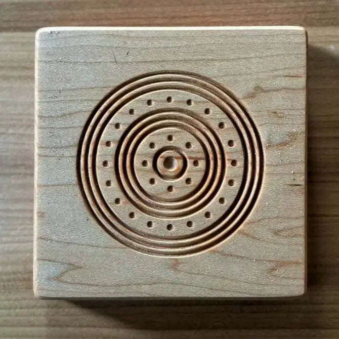 A square maple wood ravioli mold with a central circular shape and small holes on the surface, accompanied by a mini pin for pressing.