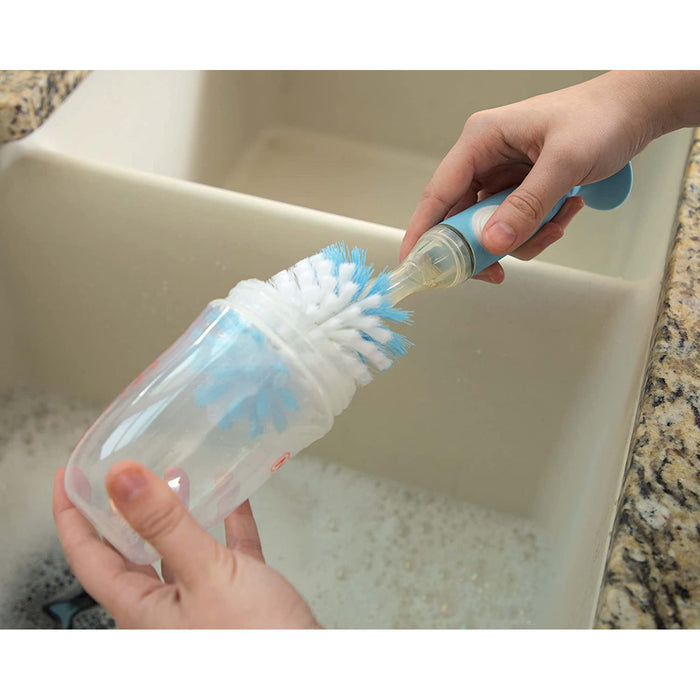 A person uses the Easy Clean Soap Dispensing Brush with Suction Base to clean a clear plastic bottle over a sink filled with soapy water.