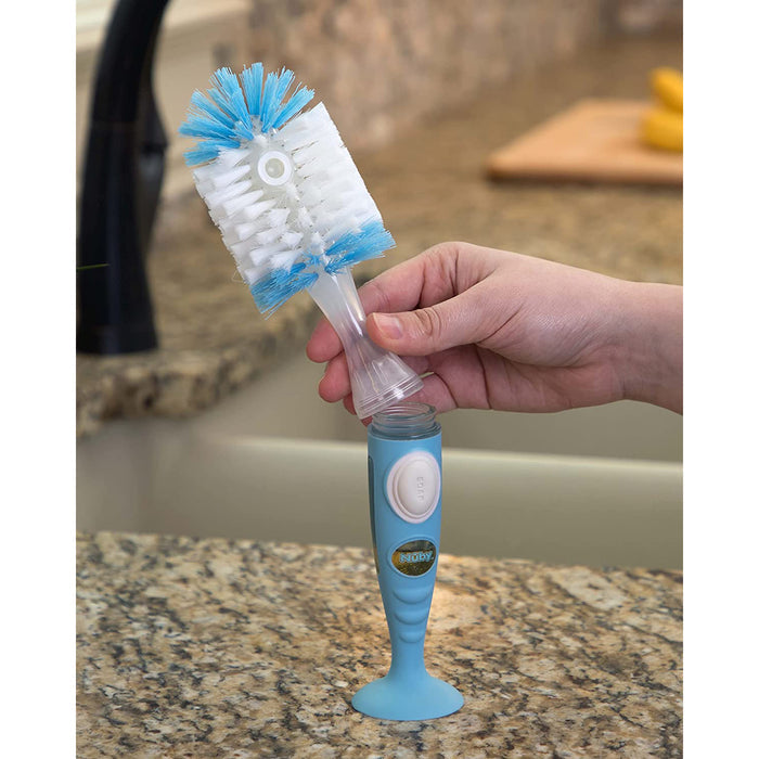 A hand holds the Easy Clean Soap Dispensing Brush with Suction Base, featuring blue and white bristles and a built-in soap dispenser, above a granite kitchen countertop near a sink.