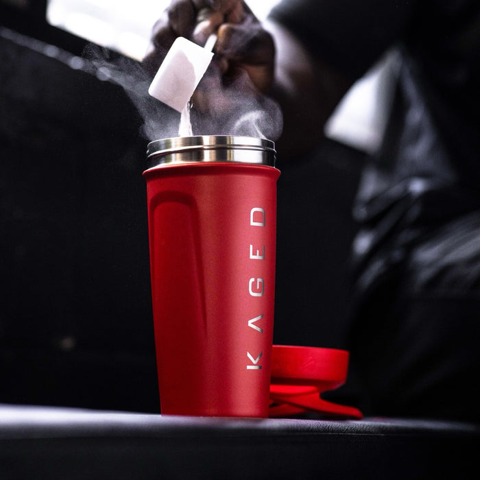 Three-quarter view of Elite Strada red shaker bottle showing KAGED logo, open stainless rim as protein powder is poured