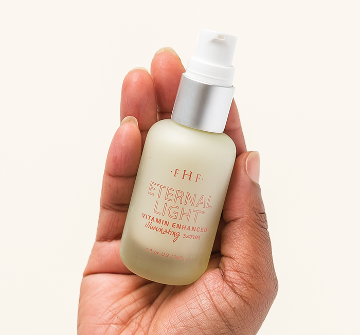 Hand holding a frosted glass bottle of Eternal Light® Vitamin Enhanced Illuminating Serum | Farmhouse Fresh with a white pump dispenser against a neutral background.