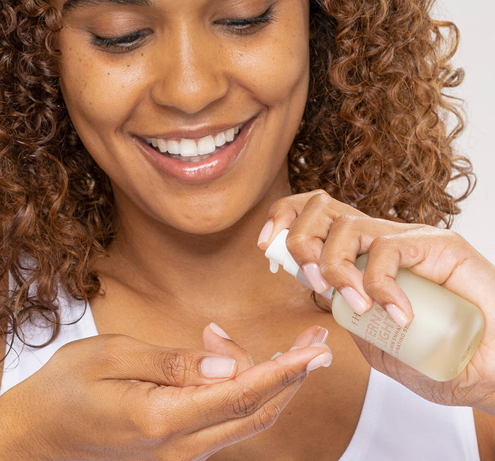 A smiling woman with curly hair dispensing Eternal Light® Vitamin Enhanced Illuminating Serum | Farmhouse Fresh onto her hand, demonstrating skincare use.