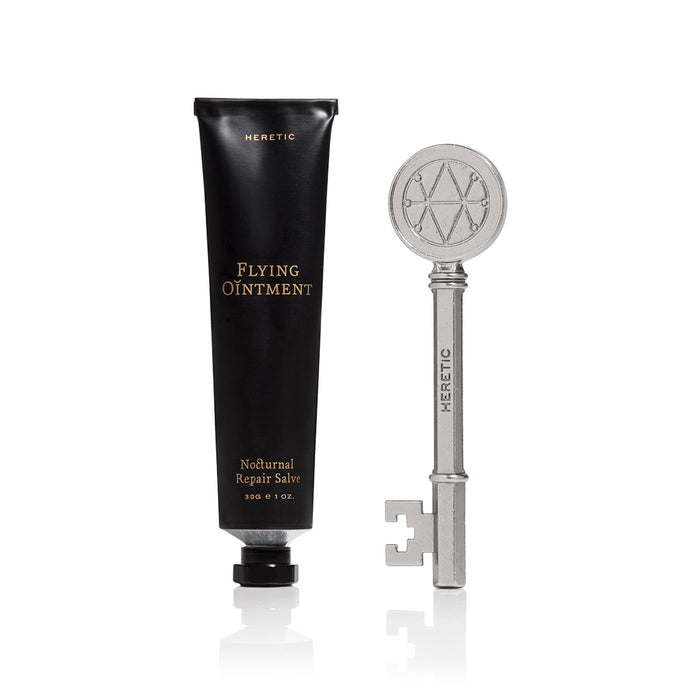 A black squeeze tube labeled 'HERETIC Flying Ointment' stands next to a silver key with a circular emblem on the head.