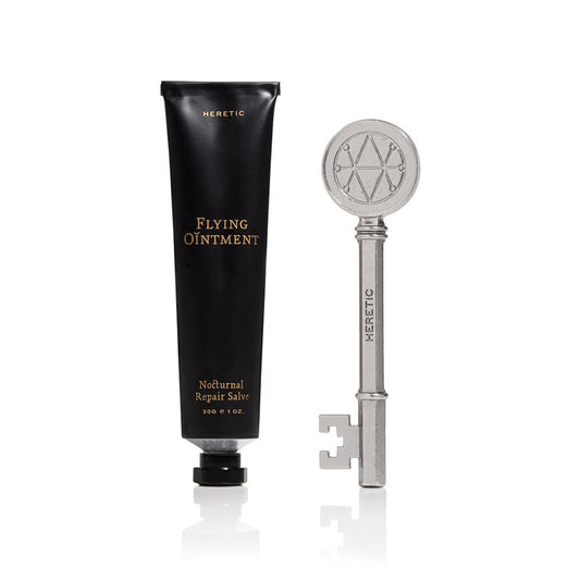 A black squeeze tube labeled 'HERETIC Flying Ointment' stands next to a silver key with a circular emblem on the head.