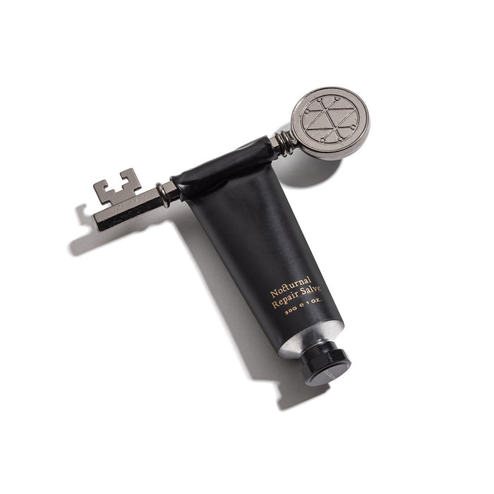 A black hand cream tube with gold lettering reading 'Nocturnal Repair Salve' and a silver key to assist with squeezing product out.