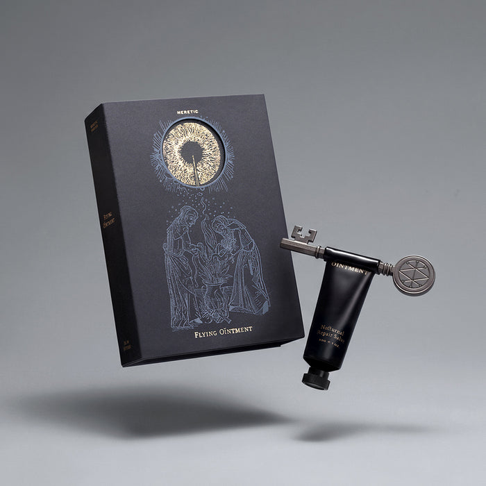 A black presentation box with a circular window showing a gold-toned circular design, alongside a black hand and cream tube and a silver key, all floating against a neutral gray background.