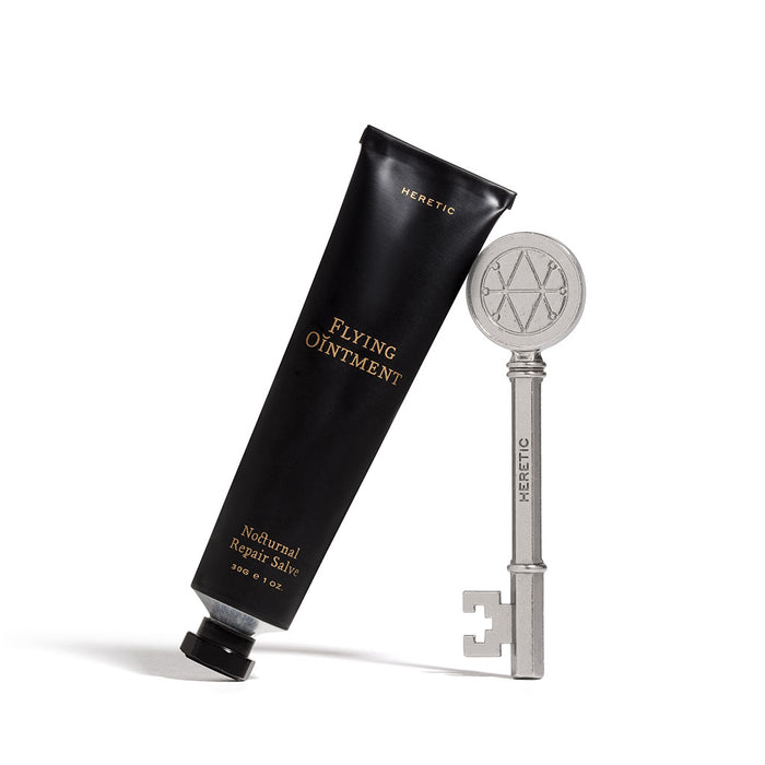 A black squeeze tube labeled 'HERETIC FLYING OINTMENT' stands beside a silver key engraved with the same brand name and a pentagram-like symbol, on a clean white background.