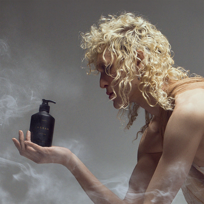 A model with voluminous curly blonde hair holds a black Haxan lotion bottle in their hand, surrounded by a dramatic misty atmosphere.