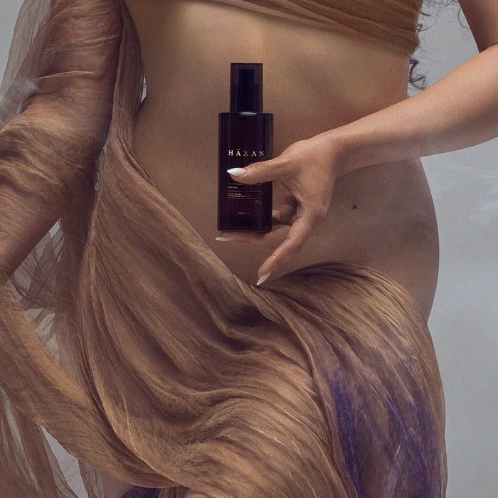 An ethereal woman in a flowing sheer brown dress holds a Haxan body oil product.