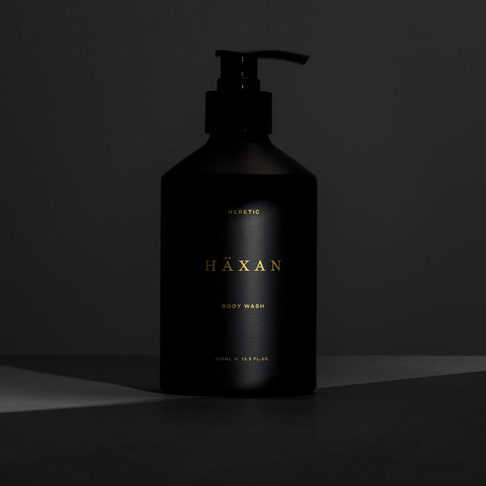 A matte black pump bottle of Häxan body wash with gold lettering, set against a dark gray background with dramatic side lighting.