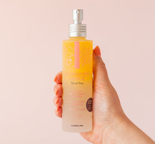 Hand holding a spray bottle of Illumination Juice® Facial Tonic – Illuminating Facial Toner | Farmhouse Fresh with a light pink background, highlighting skincare and wellness.