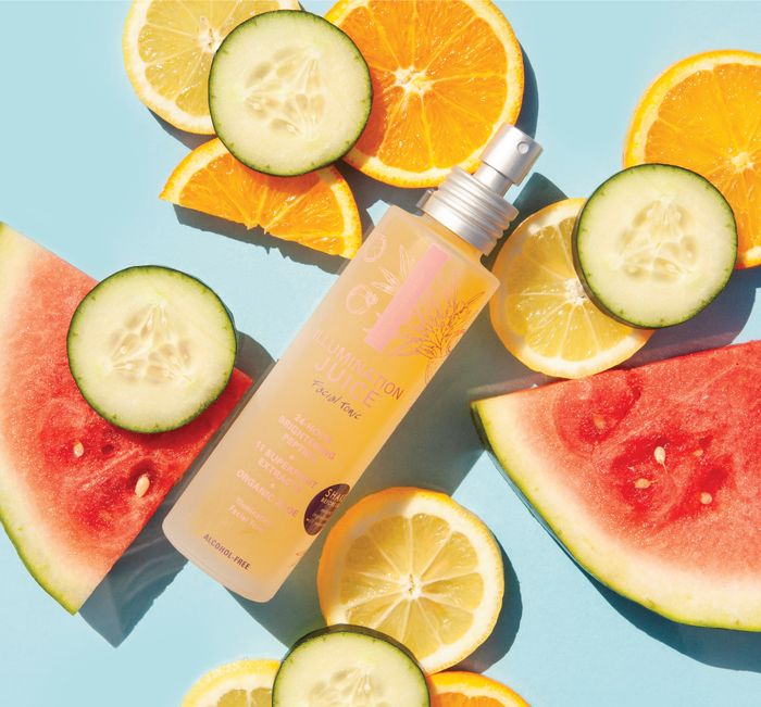 Bottle of Illumination Juice® Facial Tonic – Illuminating Facial Toner | Farmhouse Fresh surrounded by fresh citrus slices, cucumber, and watermelon on a blue background for skincare and wellness.