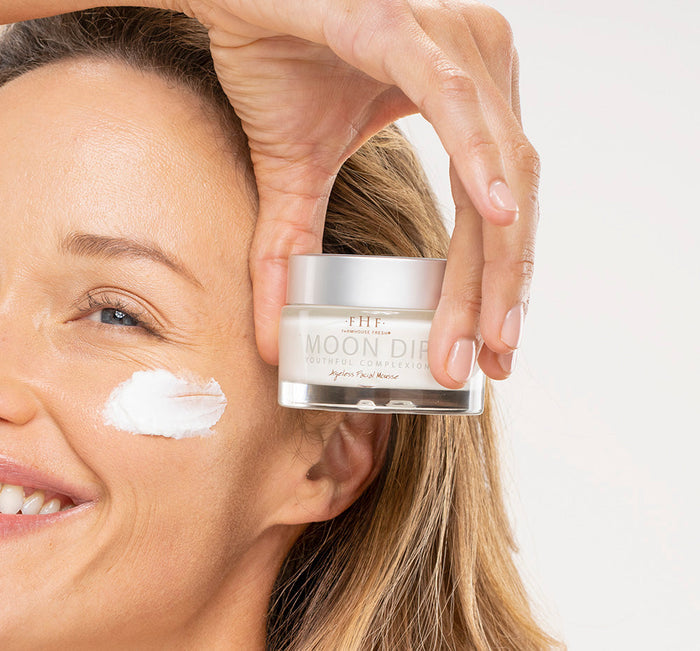 Smiling woman with glowing skin holds a jar of Moon Dip® Youthful Complexion Ageless Facial Mousse with Peptides + Retinol | Farmhouse Fresh while showing a dab of the cream on her cheek in a skincare setting.