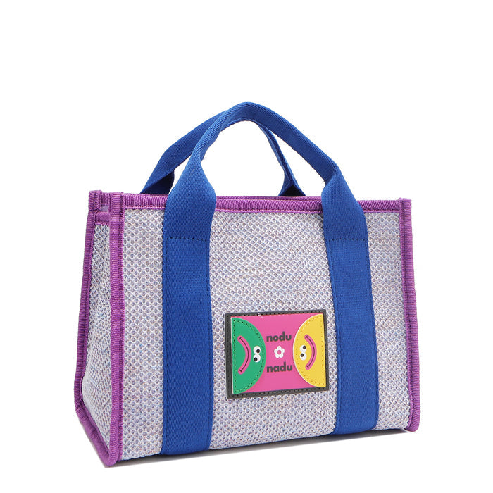 Front angle view of the Nodu Nadu small Marley tote bag in lavender.  Featuring dual top handles with a 3.5" drop and adorned with a logo stitched nodu nadu patch with a smiley face and sad face. Metallic shimmering light lavender canvas with bold electric blue strap accents complimented with a vibrant purple trim.