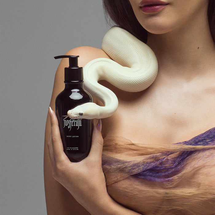 A woman holds a black pump bottle against her shoulder, with a pale snake draped over her shoulder and a purple-and-tan garment visible. The background is a plain light gray studio backdrop.