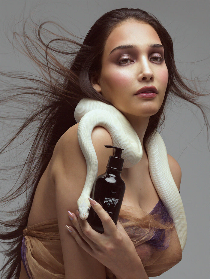 A woman with long dark hair poses with a white snake draped over her shoulders and holds a black bottle.