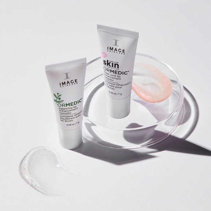 Two tubes of ORMEDIC lip enhancement complex | IMAGE Skincare displayed on a clear round dish with clear and pink gel samples nearby on a white surface.
