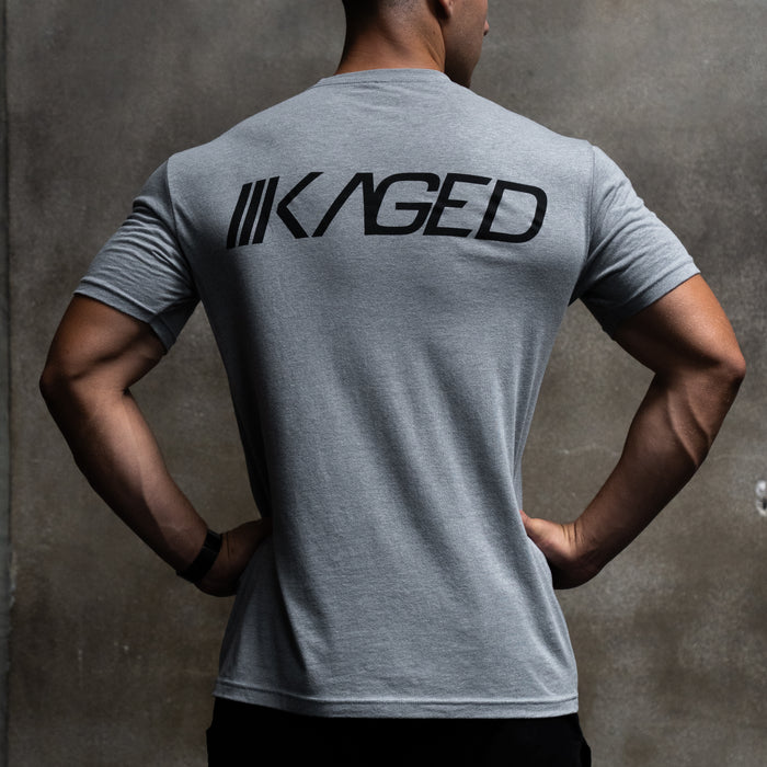 Back view of Slant Tee in gray showcasing large black KAGED logo across upper back with concrete wall background #color_gray