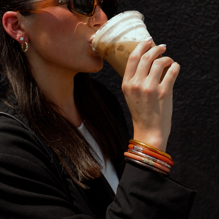 A close-up lifestyle shot of a woman wearing stacked bangles, sunglasses, and small hoop earrings while drinking from an iced coffee. The bangles feature a mix of gold, orange, and brown tones with some silver engraved details.