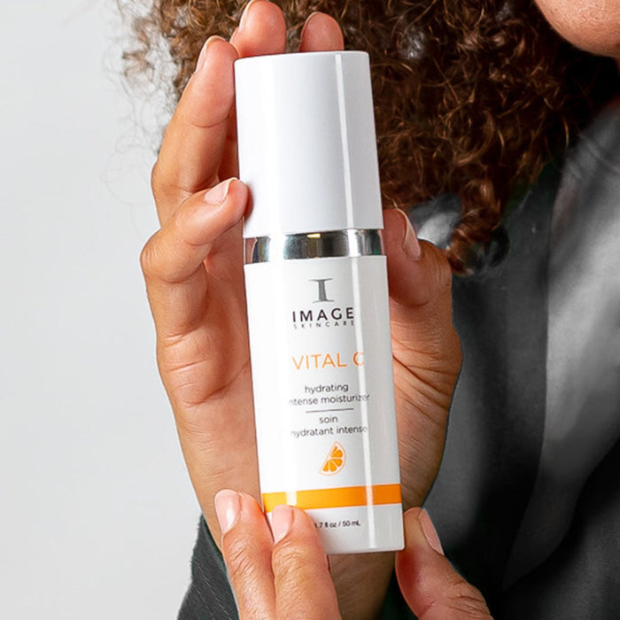 Hand holding a bottle of VITAL C hydrating intense moisturizer | IMAGE Skincare, a white skincare product container with orange accents, promoting hydration and skincare wellness.