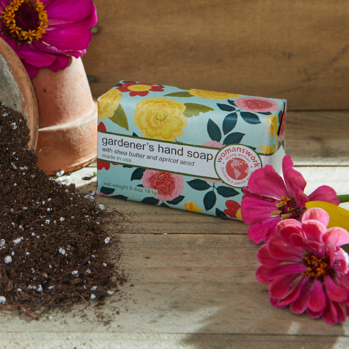 Bar of 'gardener's hand soap' with floral packaging on a wooden surface surrounded by pink flowers and soil.